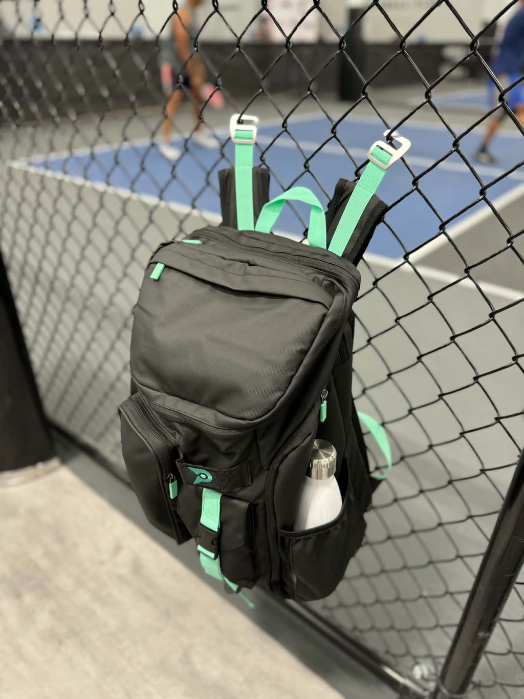 PVT Pickleball ARC bag in black waterproof nylon with mint green accents hanging on a court fence with a water bottle
